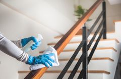 Person wearing blue gloves, cleaning a wooden handrail with spray and a white cloth. Stairs in background.