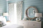 Bedroom with blue shiplap walls, white dresser, large shell-framed mirror, and patterned curtains.