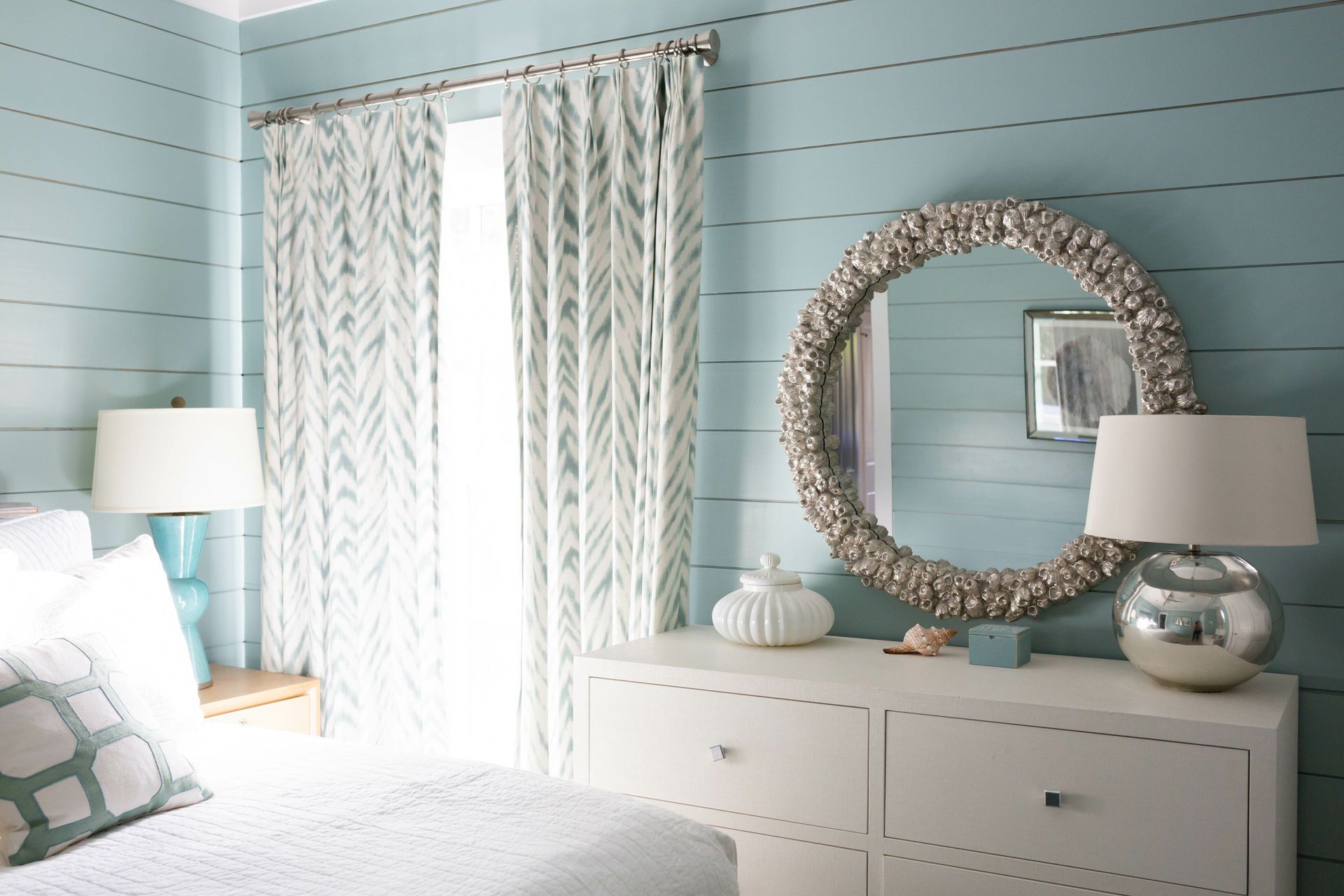 Bedroom with blue shiplap walls, white dresser, large shell-framed mirror, and patterned curtains.