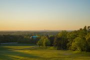 Golden sunset over a field, with trees in the foreground and buildings in the hazy distance.