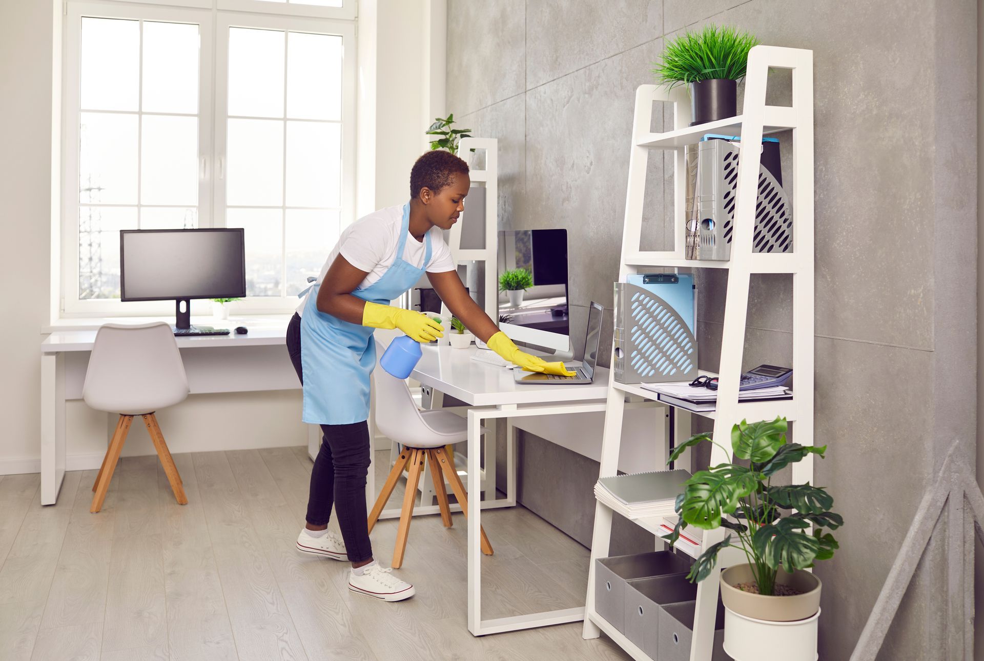 Person wearing gloves and apron cleaning an office desk with a spray bottle and cloth.