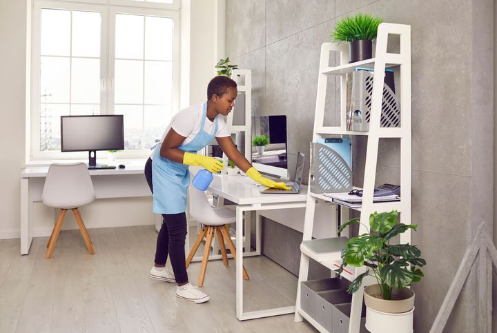 Person wearing gloves and apron cleaning an office desk with a spray bottle and cloth.