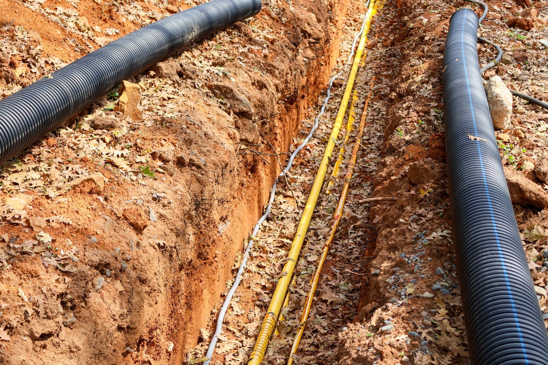 Trench in reddish-brown soil with yellow and white cables, and two black corrugated pipes.