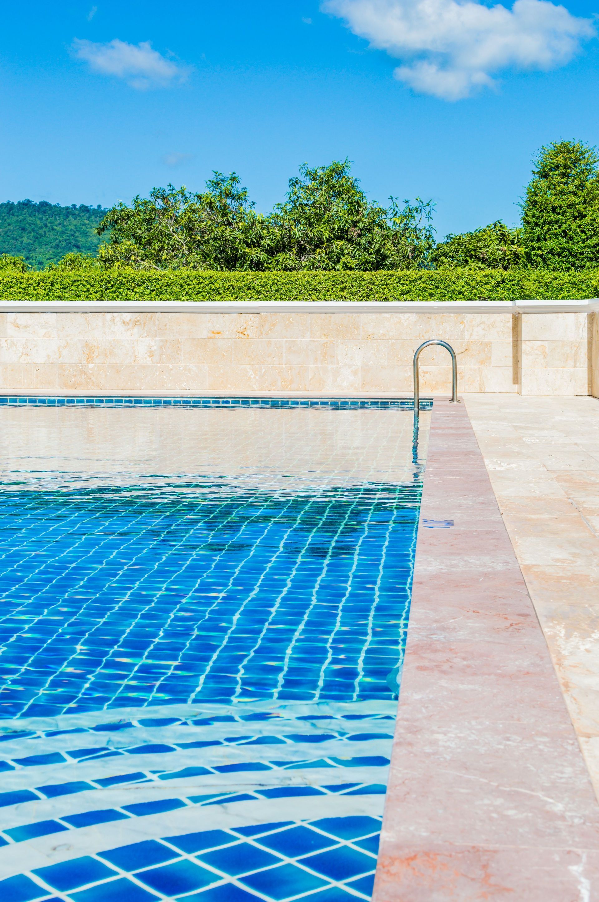 Blue-tiled swimming pool with clear water and a ladder, bordered by a stone deck and greenery under a blue sky