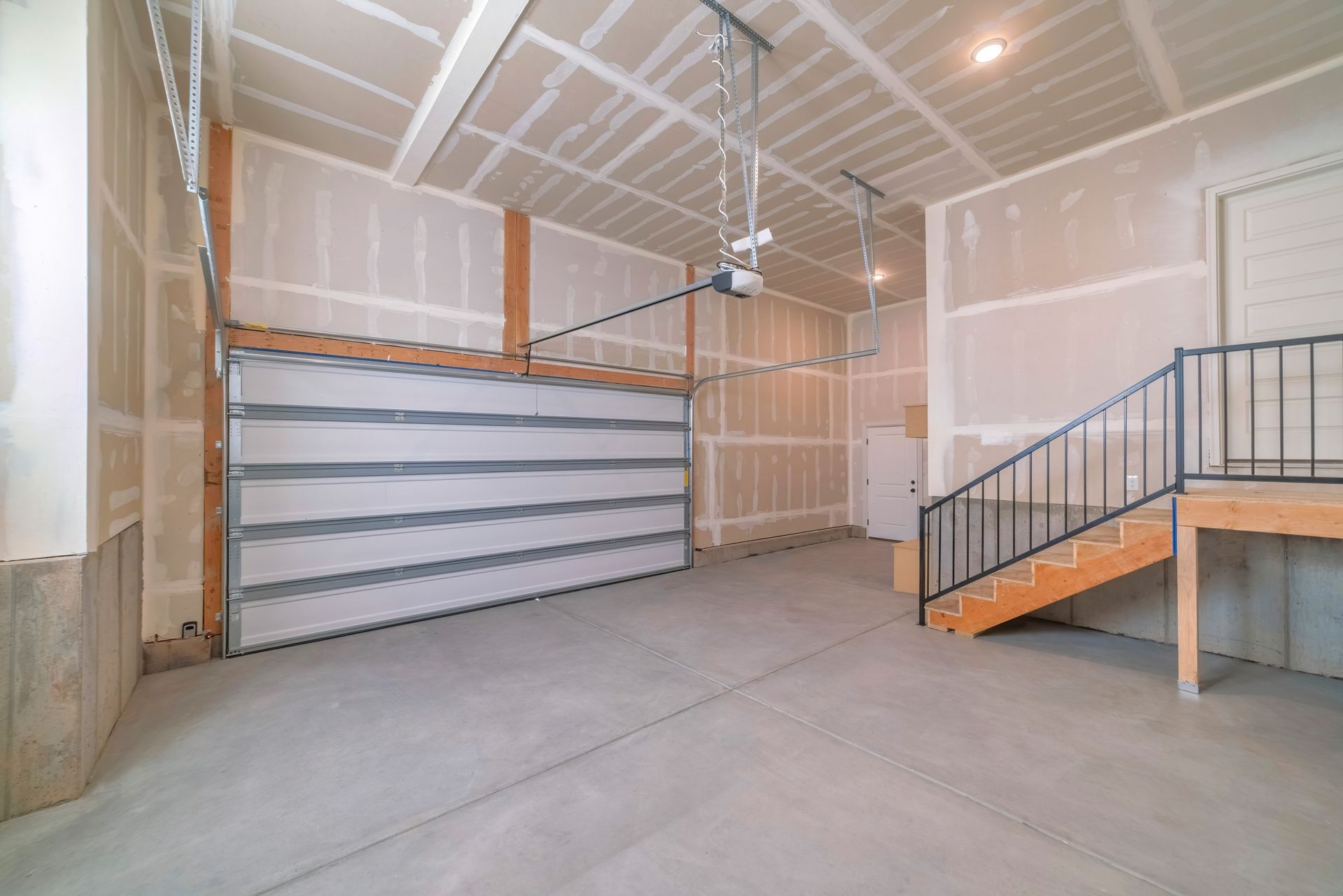 Empty garage interior with open garage door, concrete floor, drywall walls, and staircase.
