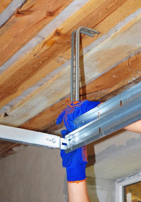 Person wearing blue work gloves installing metal framing on a wooden ceiling.