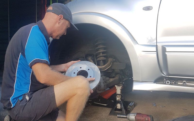 A Man Is Kneeling Down in Front of A Silver Truck Holding a Brake Disc — Automotion Mechanical In Pacific Paradise, QLD