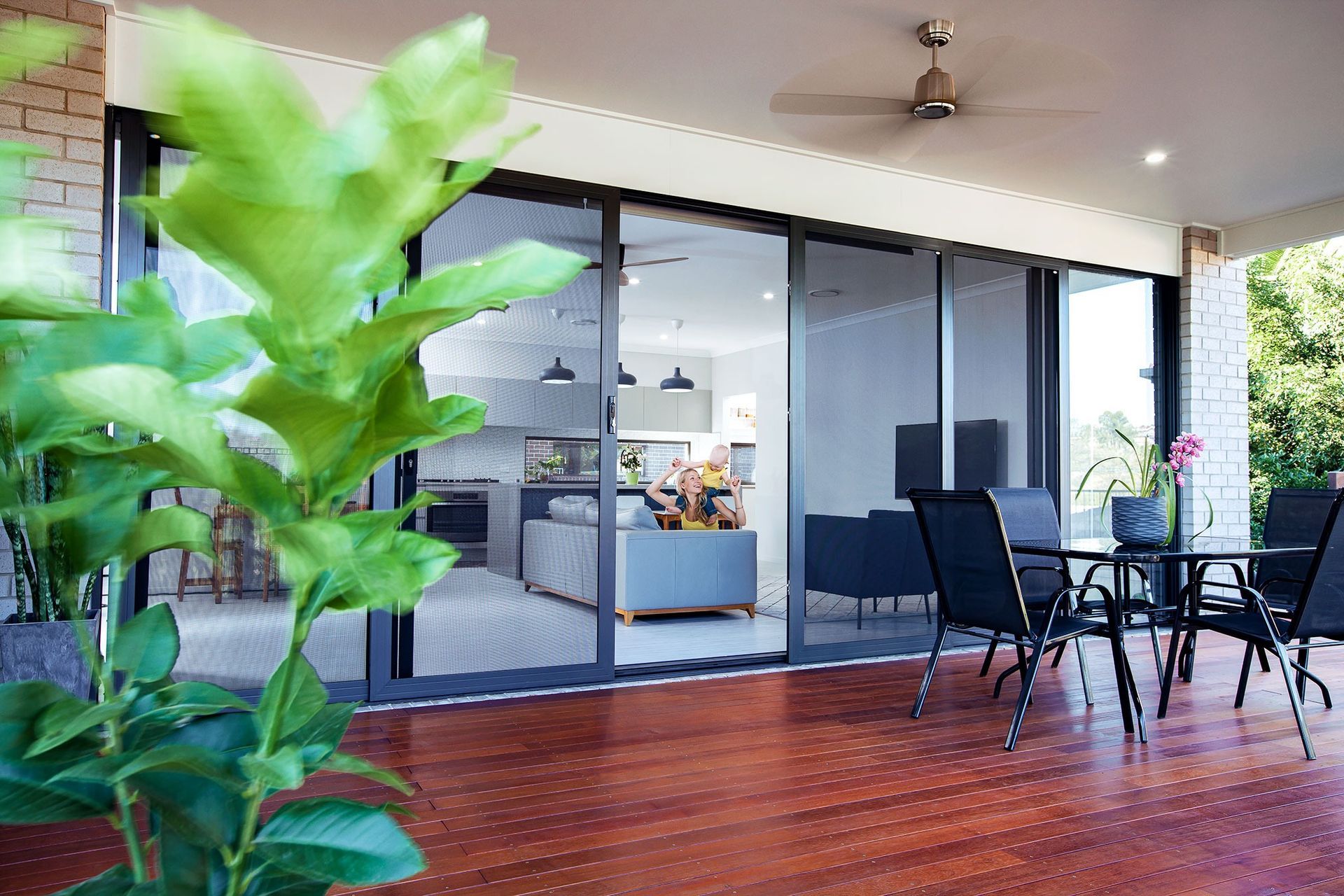 A patio with a dining table and chairs, overlooking a living room through sliding glass doors. Bright green foliage in the foreground. — Douglass Blinds & Security Screens in Southern Highlands, NSW
