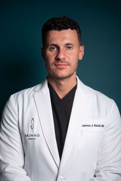 Man in white lab coat, wearing black shirt, against teal background.