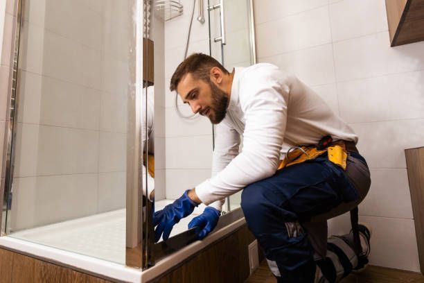 A man in a white shirt and blue gloves repairs a shower, focusing on the glass shower screen repair.