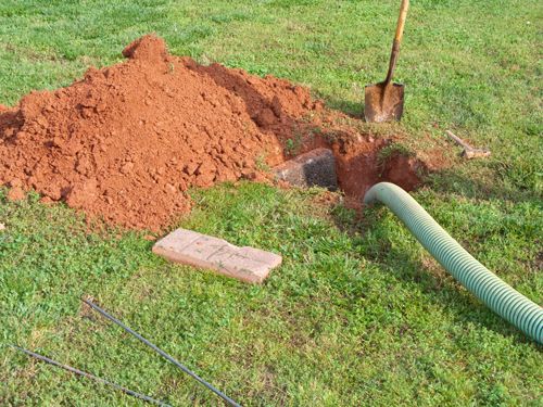 A green drainage pipe being installed in a grassy yard with a pile of dirt and a shovel nearby.