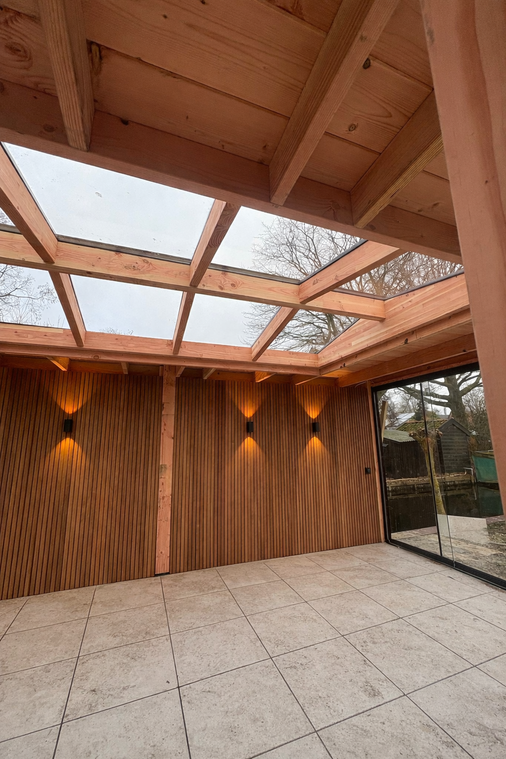 Een tuinpaviljoen met lichte houten balken, glazen dakpanelen, houten lattenwanden met sfeervolle wandlampen 