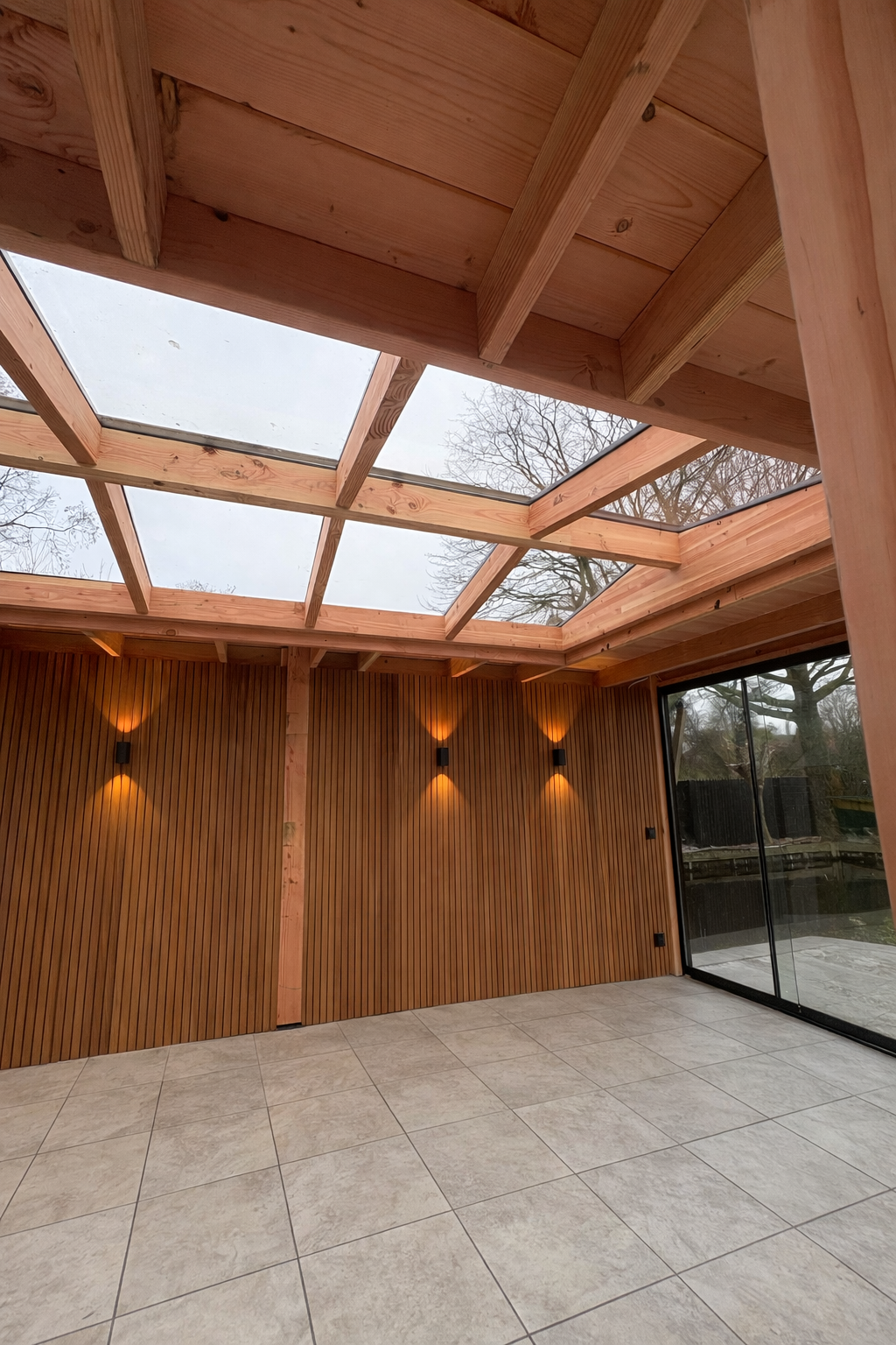 Een modern interieur van een tuinkamer met een wand van houten latten, een glazen dakraam en sfeervolle wandlampen.