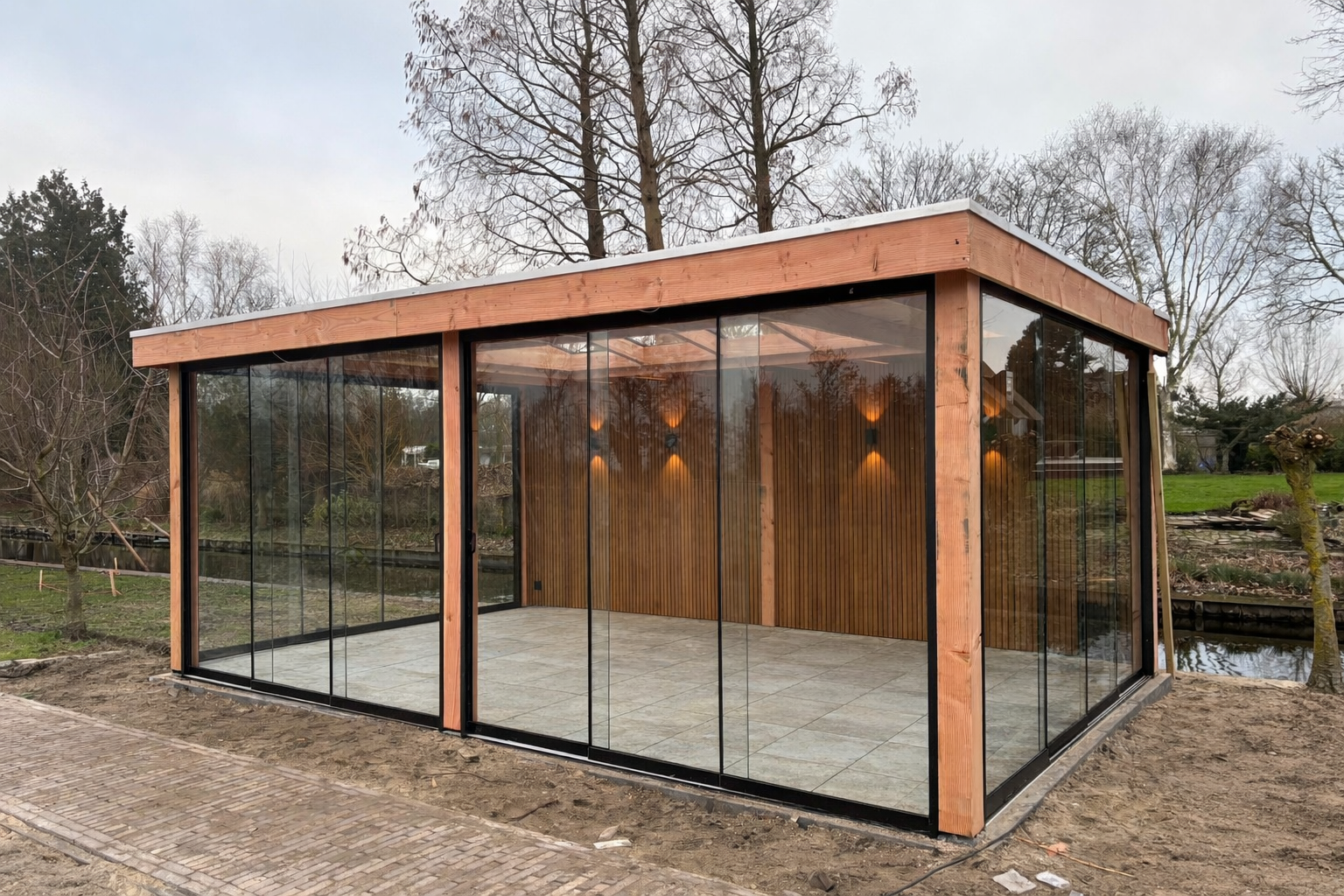 Modern houten buitenpaviljoen met glazen wanden, tegelvloer en sfeerverlichting, gelegen in een tuin aan een kanaal.