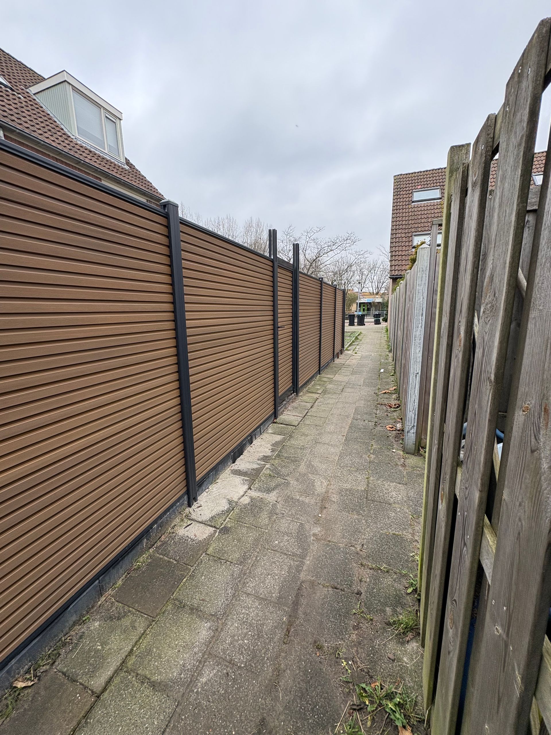 Smalle steeg tussen twee hekken; links een bruin lattenhek, rechts een verweerd houten hek.