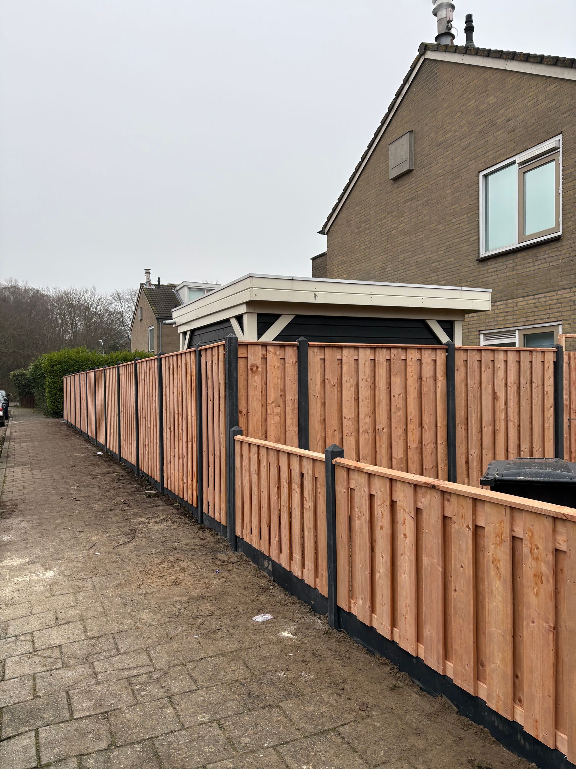 Houten hek langs een geplaveid pad, naast een bakstenen huis. Bewolkte lucht.