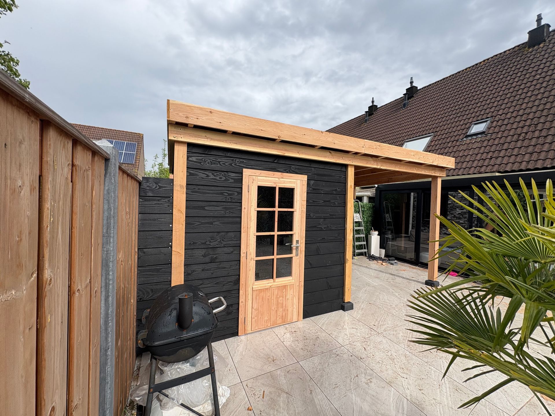 Een houten tuinschuur met een zwarte, gestructureerde buitenkant en een deur met glazen panelen, geplaatst op een terras 