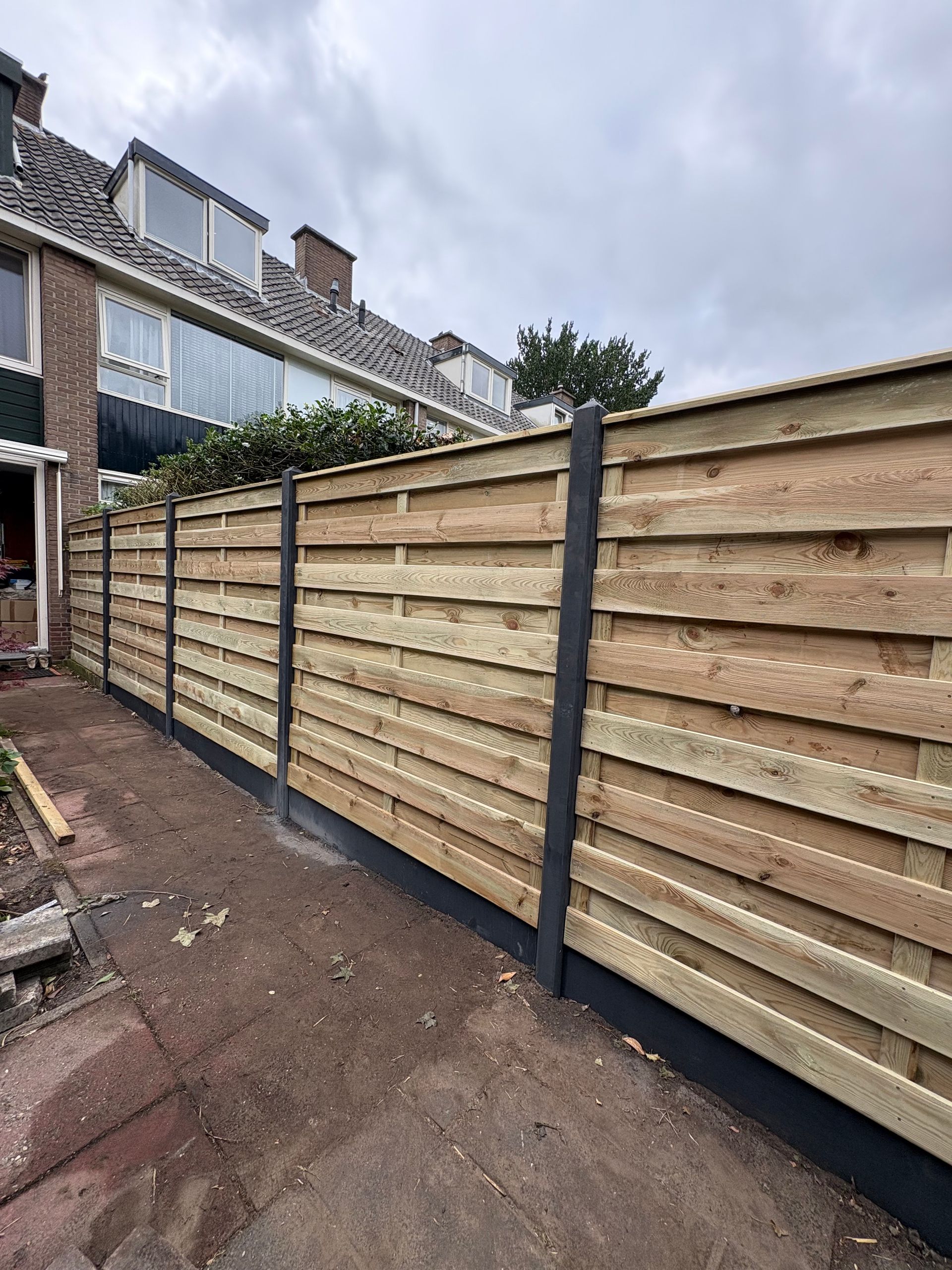 Houten schutting met donkere palen en voet, die langs de tuin voor een huis loopt.