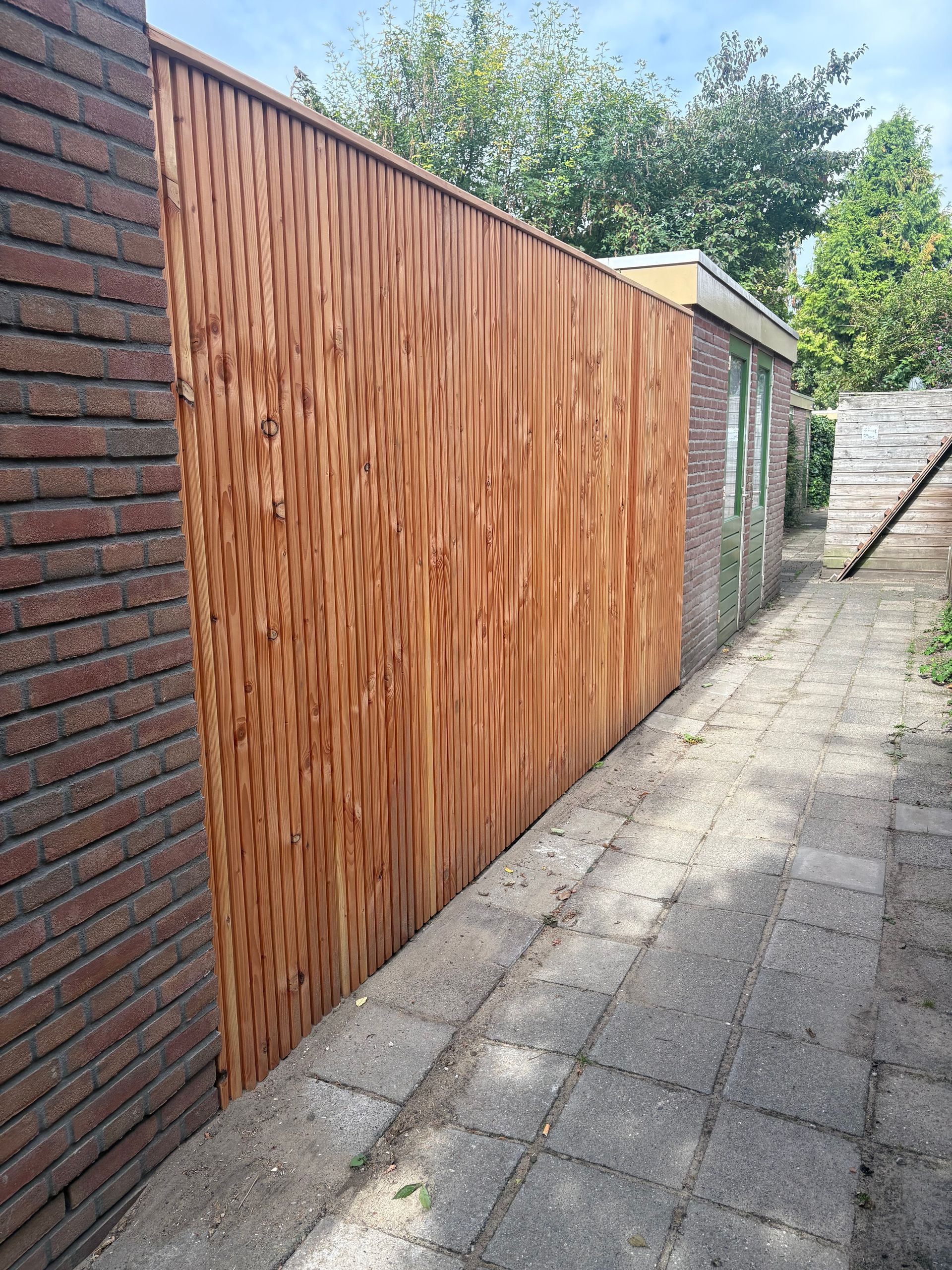 Houten schuttingpaneel bevestigd aan een bakstenen muur. Een geplaveid pad loopt erlangs.