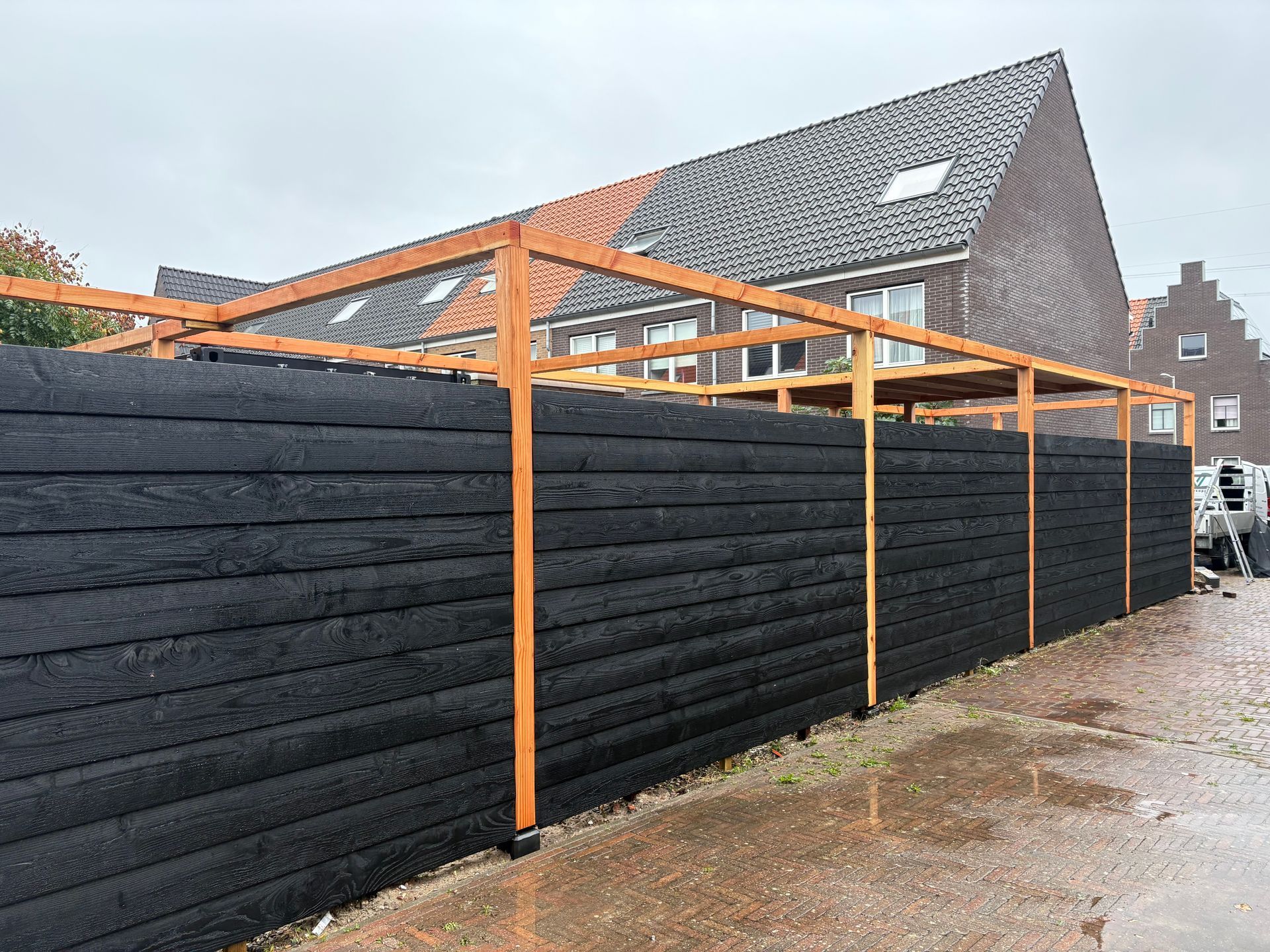 Zwarte houten schutting met een houten traliewerk, voor een bakstenen gebouw.