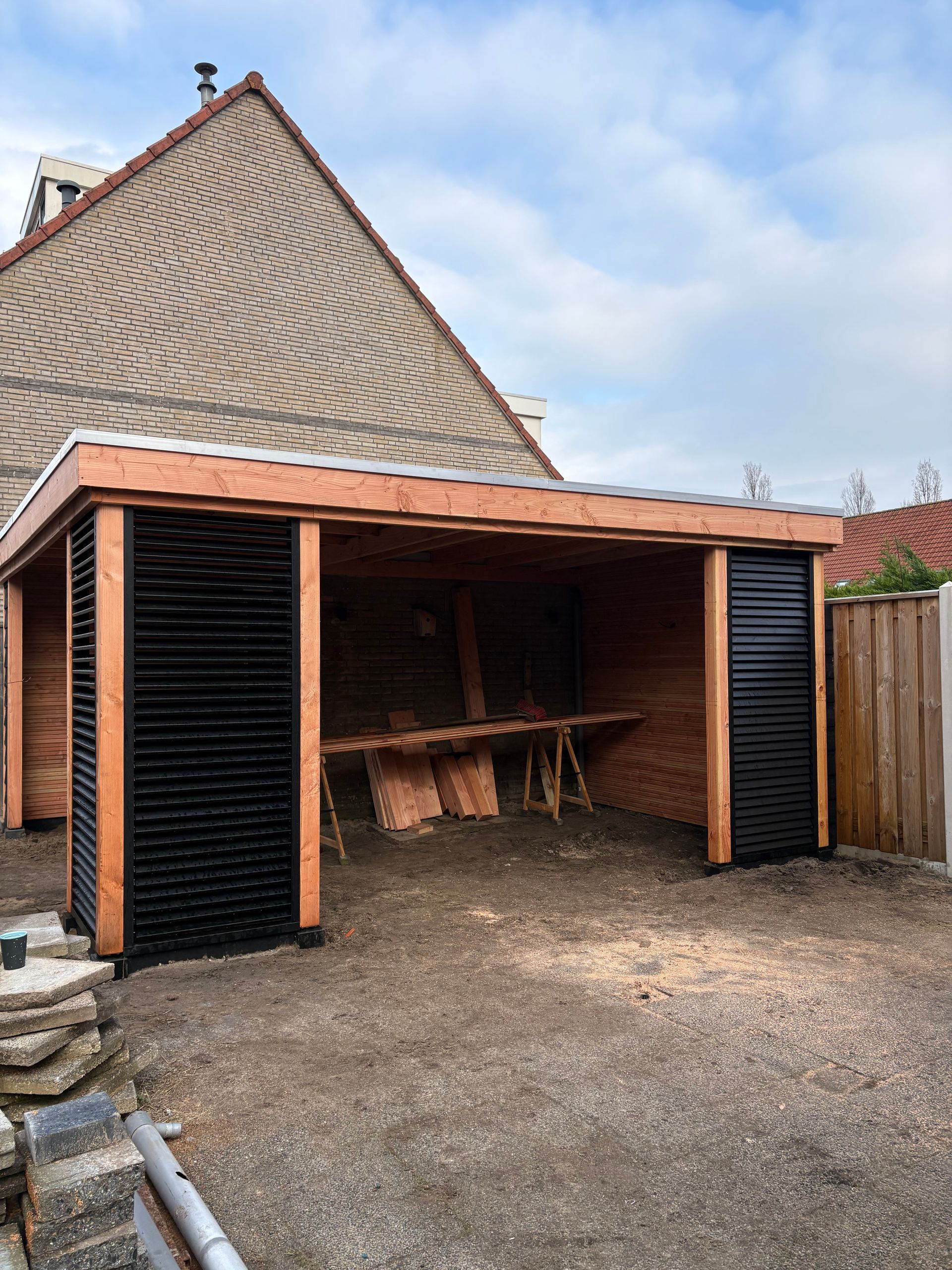 Een open houten schuur met een metalen dak en zwarte latten aan de zijkanten, gelegen in een achtertuin met een grindvloer.