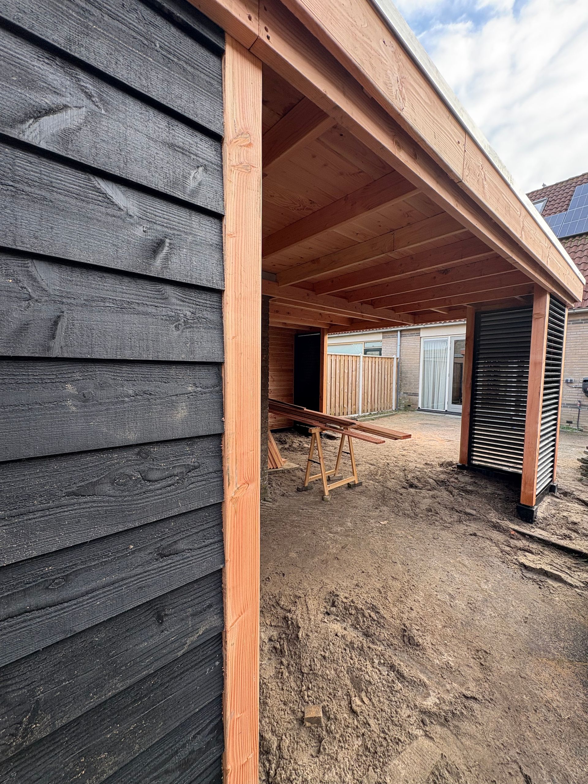 Een onafgewerkte houten constructie met zwarte houten gevelbekleding en een open buitenruimte op een onverhard terrein.