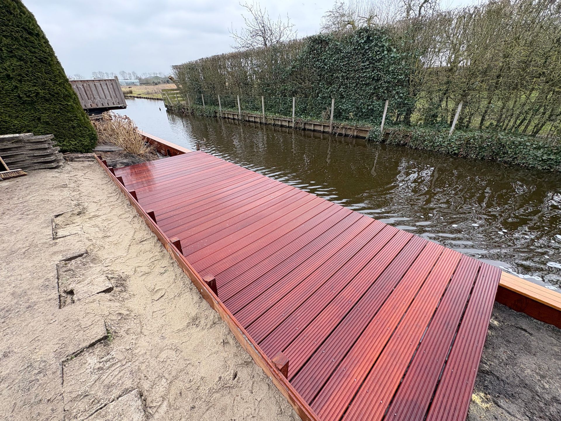 Een nieuw aangelegd, roodbruin houten terras langs een kanaal, begrensd door een zandvlakte en een groene heg.