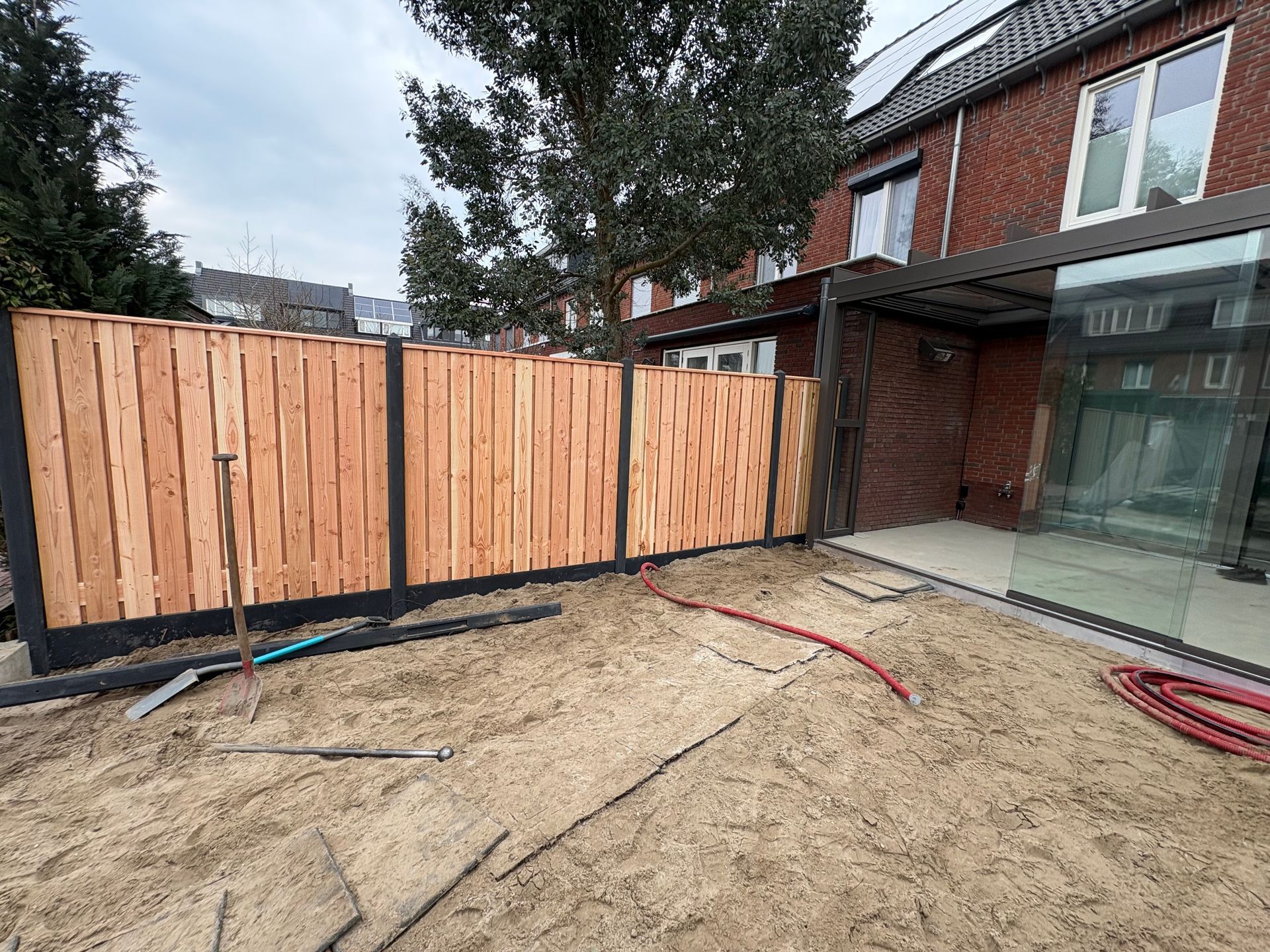 Nieuwe houten schutting en glazen terrasconstructie naast een in aanbouw zijnde woning van rode baksteen.