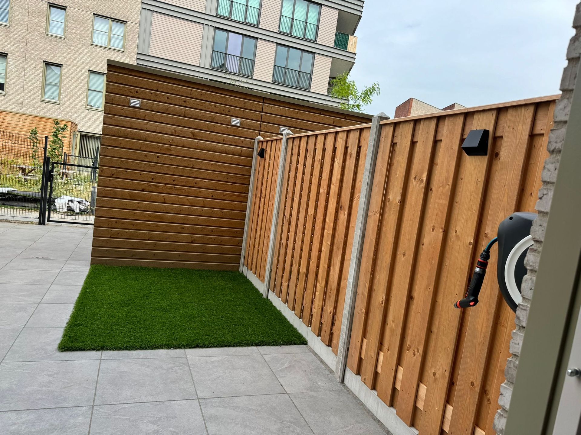 Achtertuin met een bruine houten schutting, een grijs terras en kunstgras.