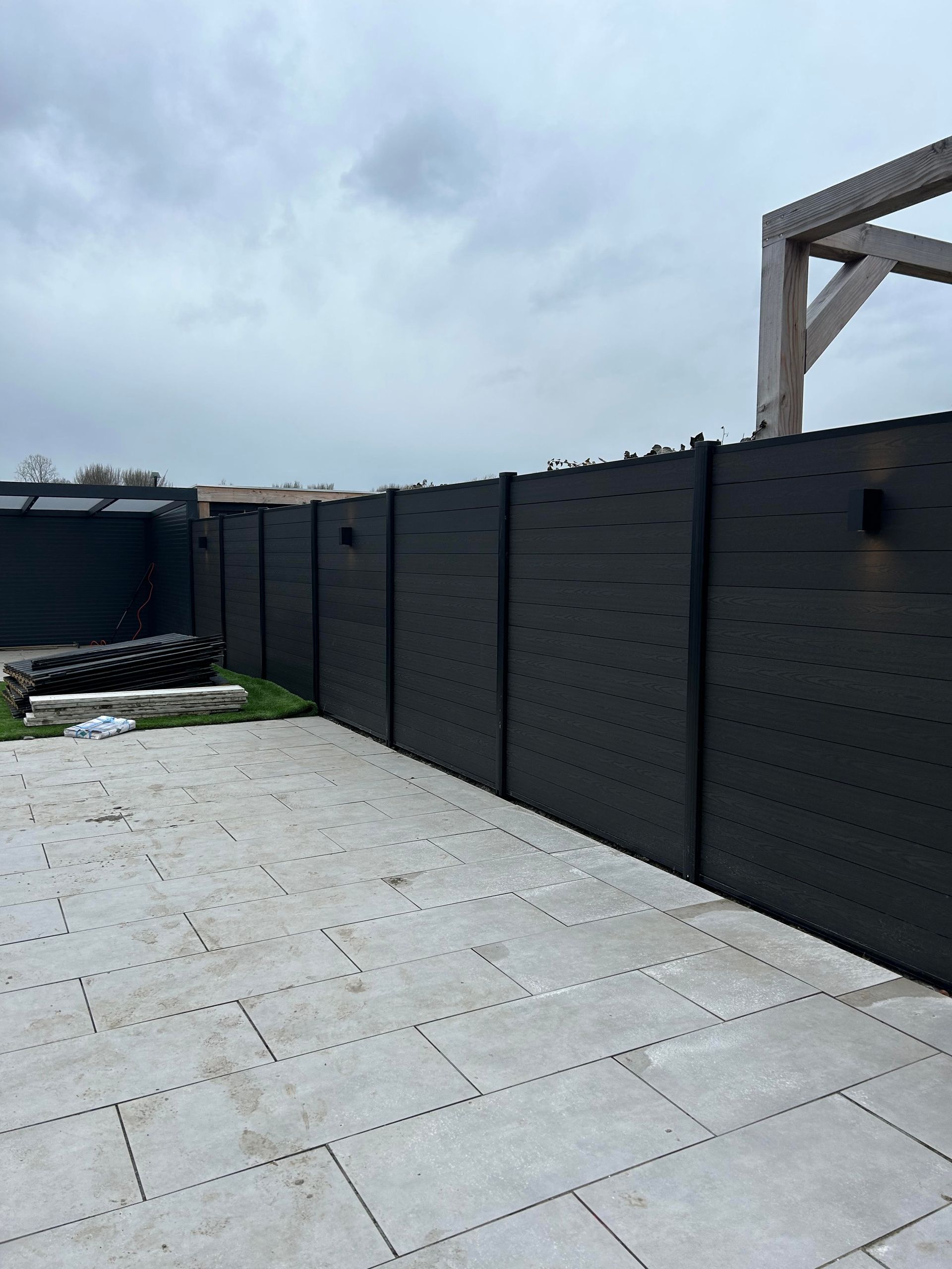 Een zwarte houten schutting langs een grijs betegeld terras onder een bewolkte hemel.