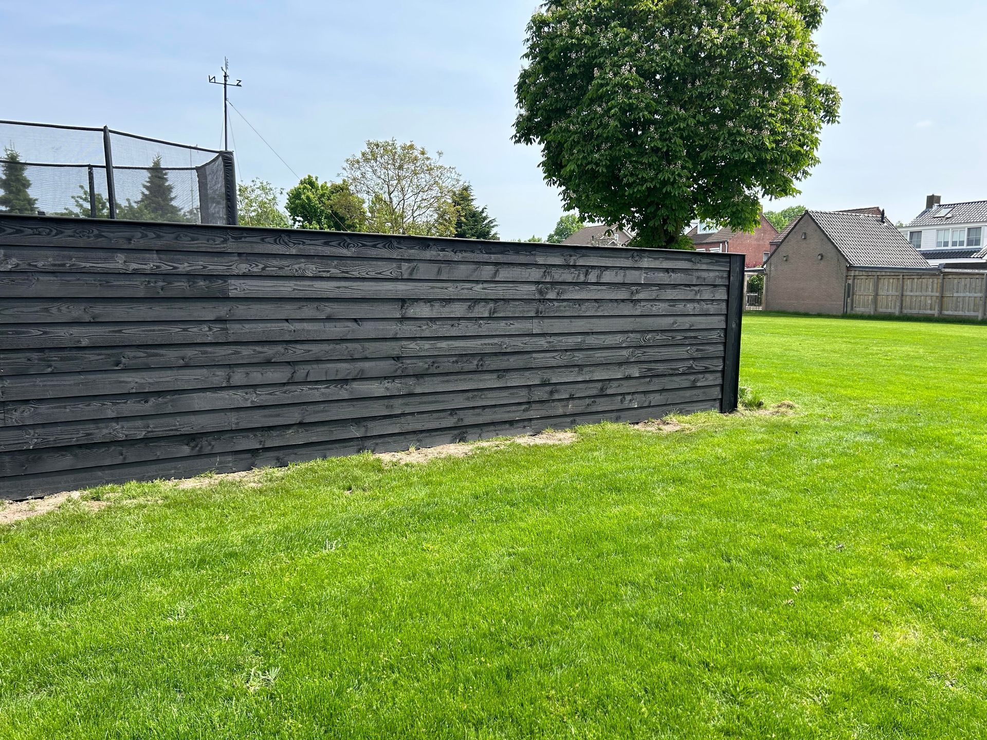 Een zwarte houten schutting in een grasveld, met een boom en huizen zichtbaar op de achtergrond.