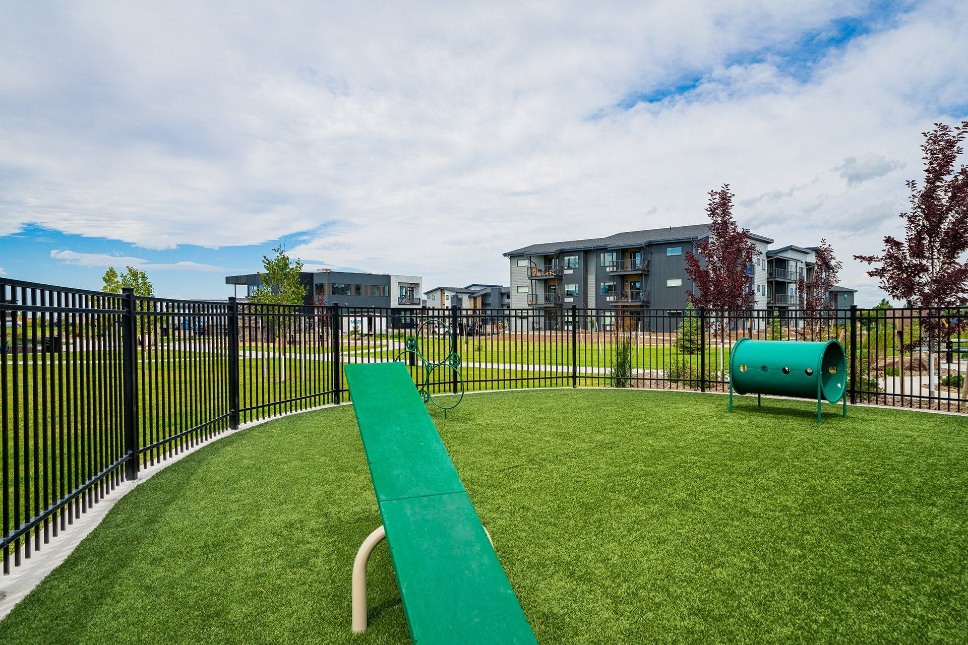 Fenced dog park with artificial turf, a seesaw, and a tunnel; apartment buildings in the background under a cloudy sky.