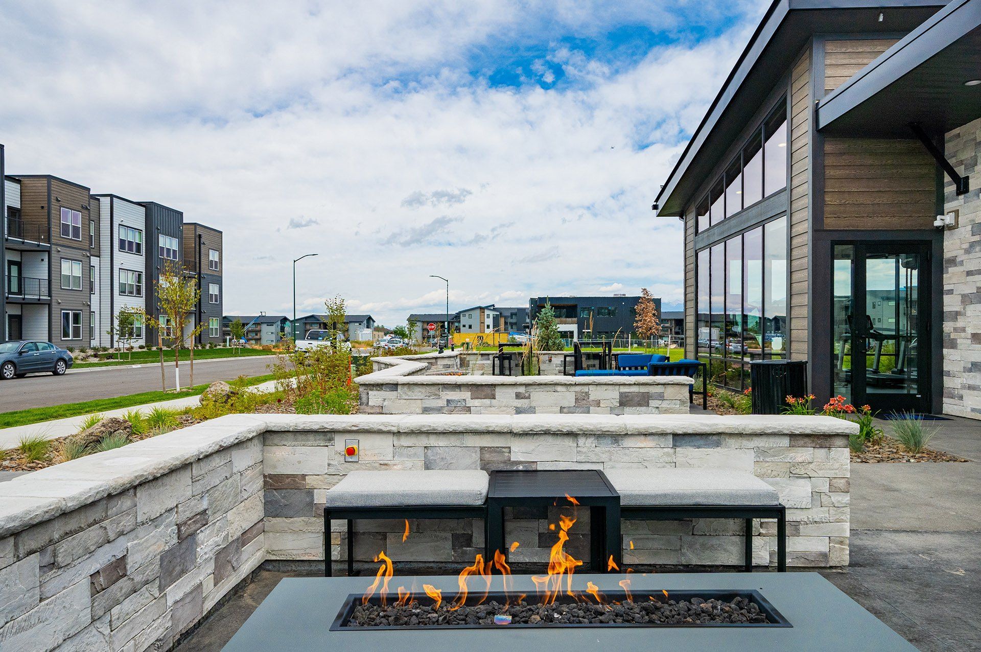 Outdoor seating area with a fire pit, modern buildings in the background, cloudy sky.