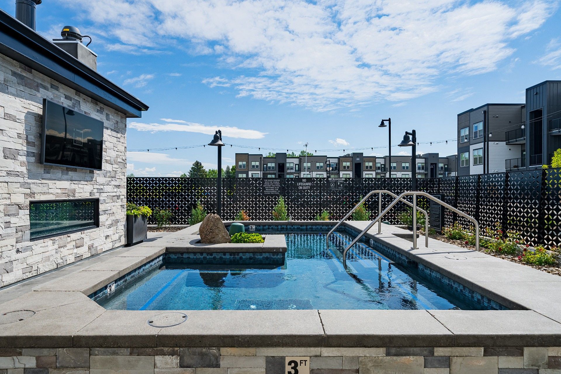 Small outdoor pool with stone surround, metal fence, and apartment buildings in the background under a blue sky.