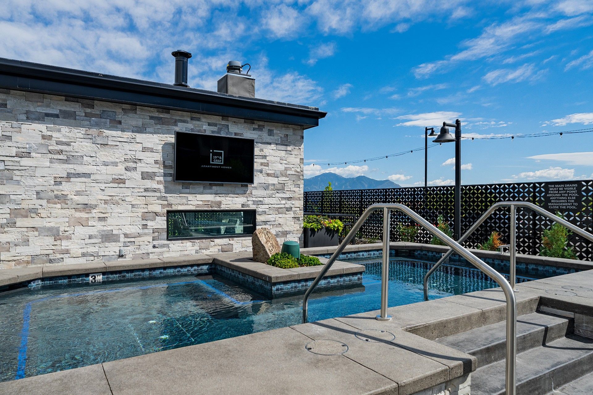 Outdoor pool with stone wall, steps, and handrails. TV mounted on the wall. Blue sky with clouds.