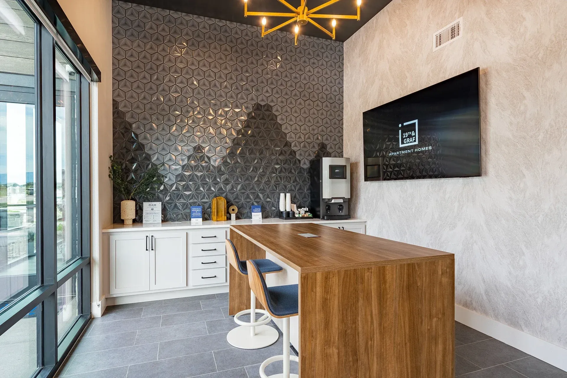 Modern break room with coffee station, textured wall, and wooden island with stools.