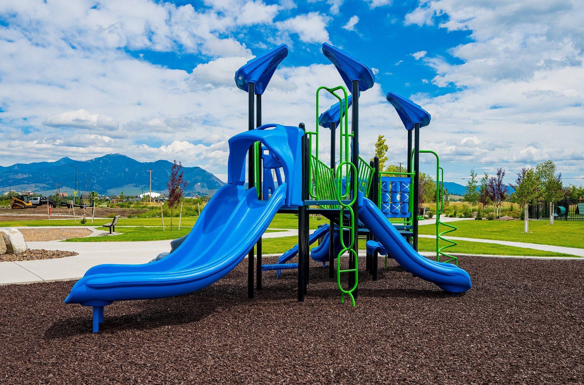 Blue and green playground slide with ladders in a park setting.