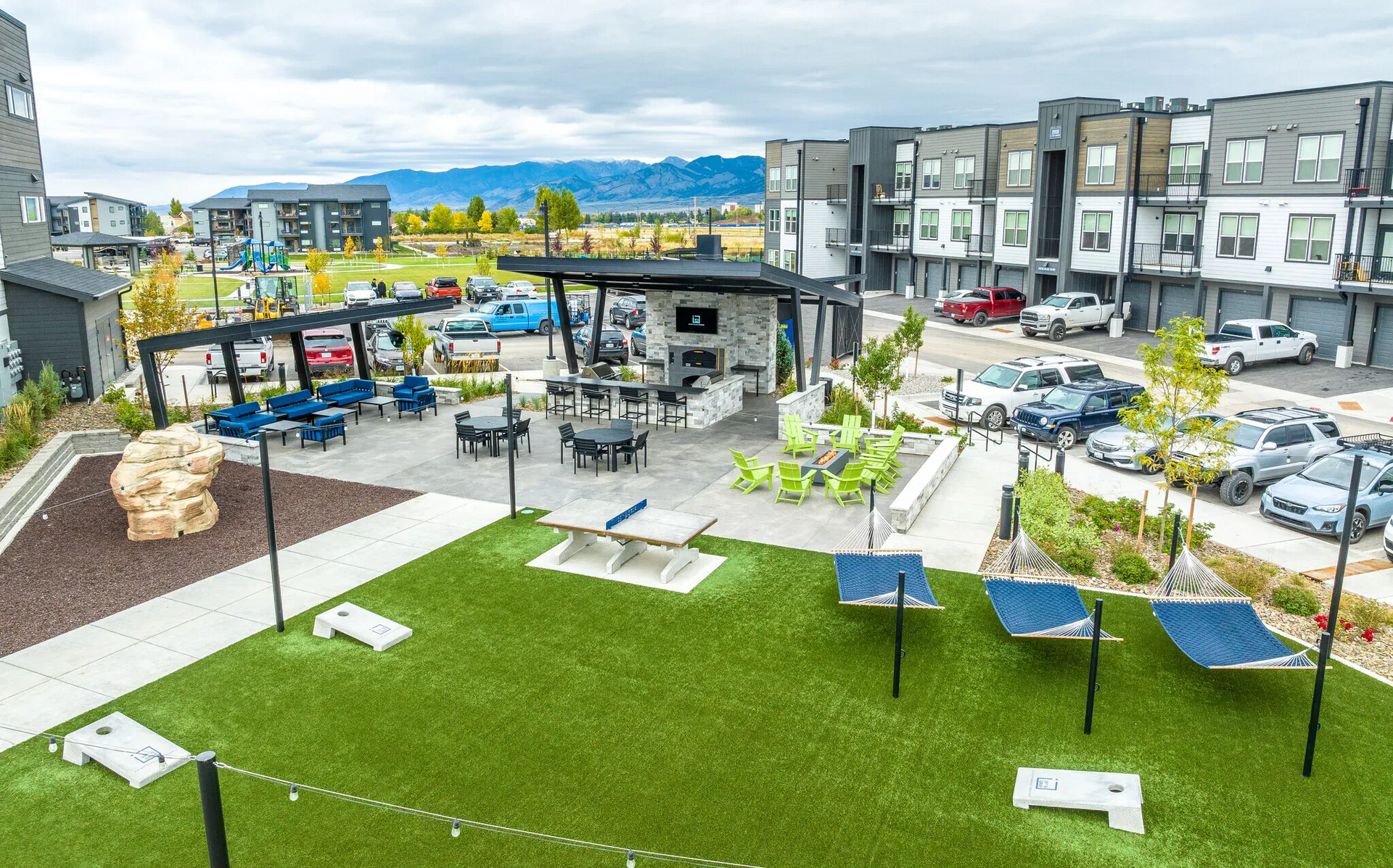 Aerial view of a residential area with buildings, roads, parking, a pool, and fields, with mountains in the background.