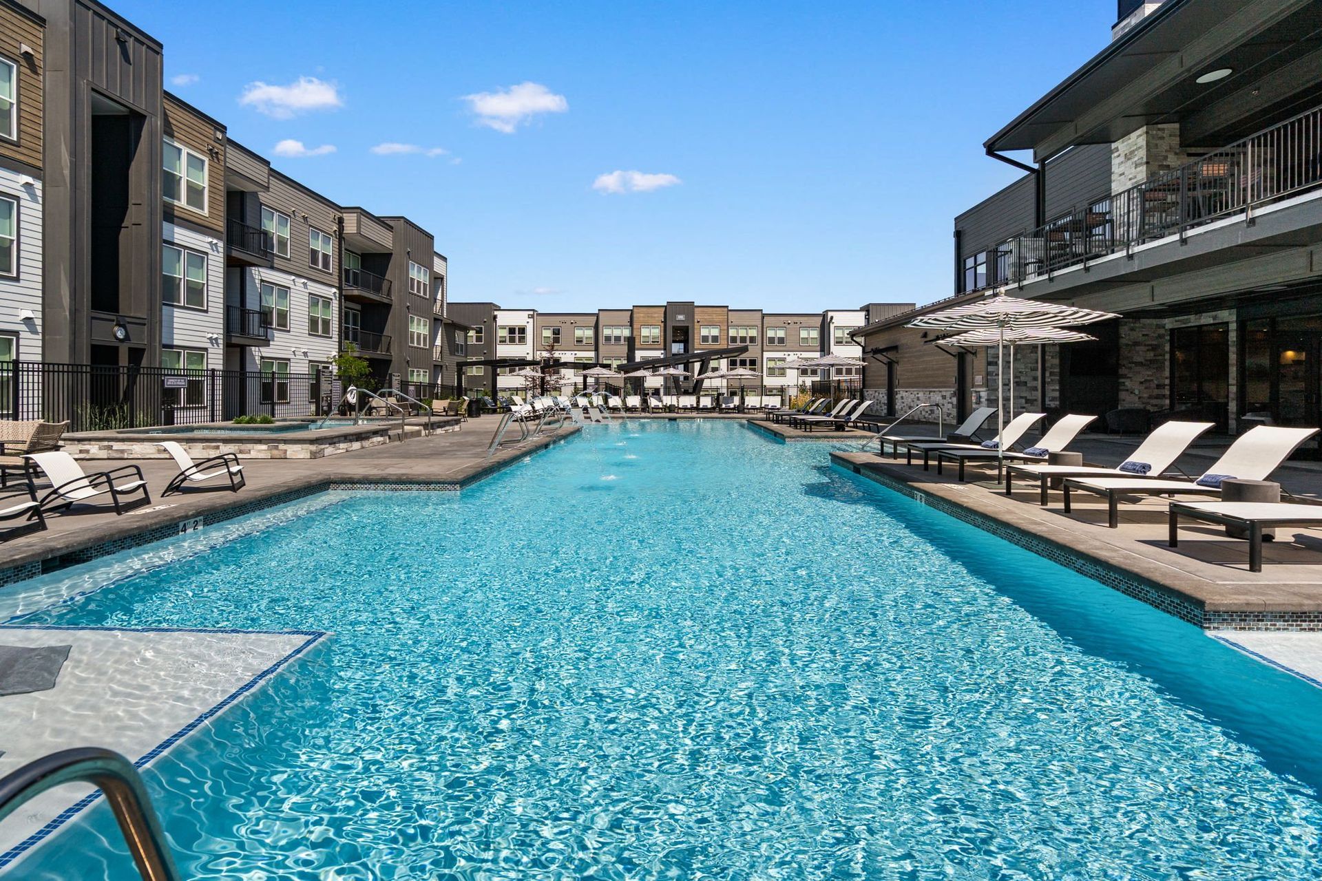 Swimming pool flanked by apartment buildings with lounge chairs. Sunny day, blue water.