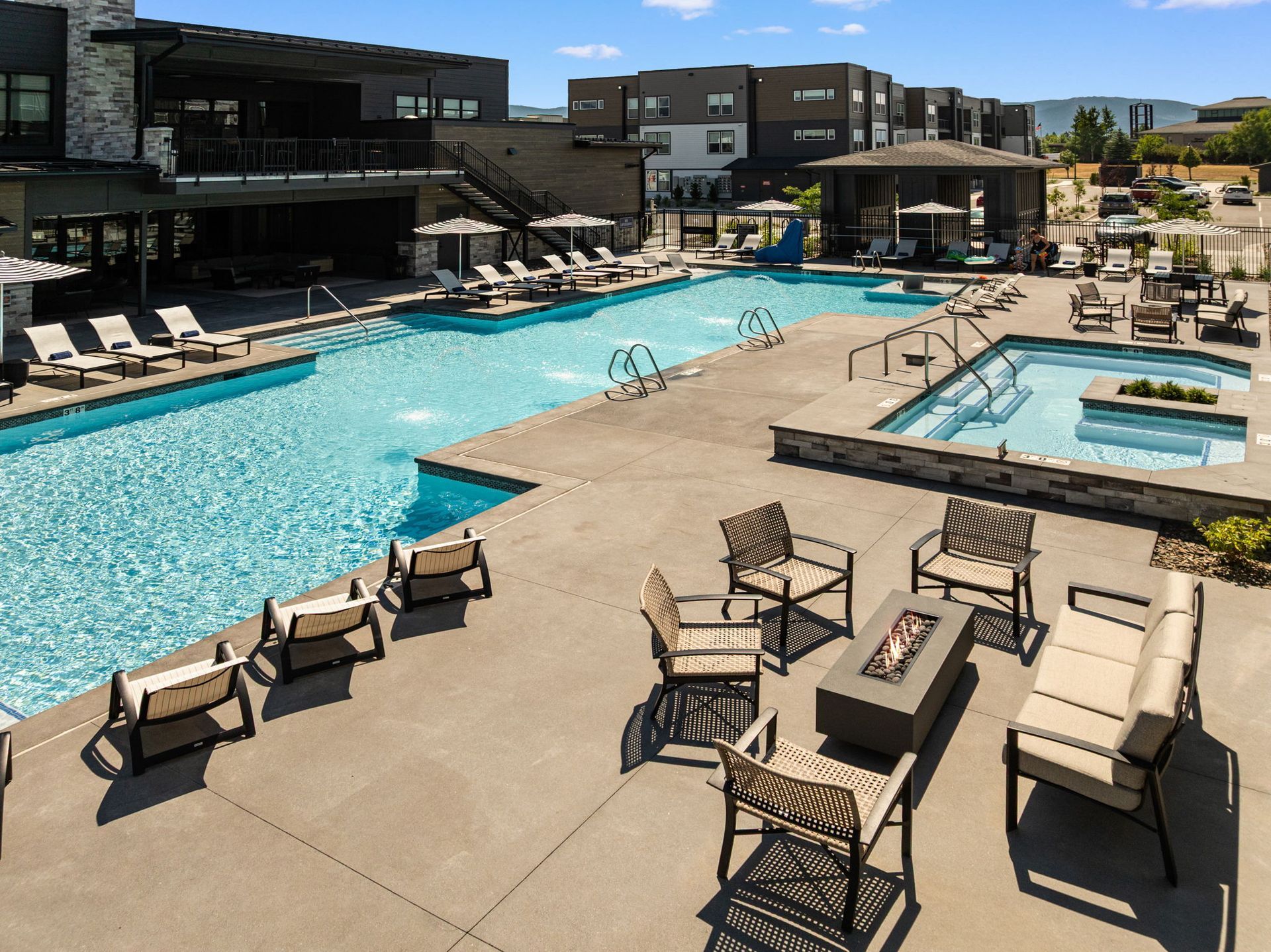Outdoor pool area with lounge chairs, fire pit, and buildings in the background on a sunny day.