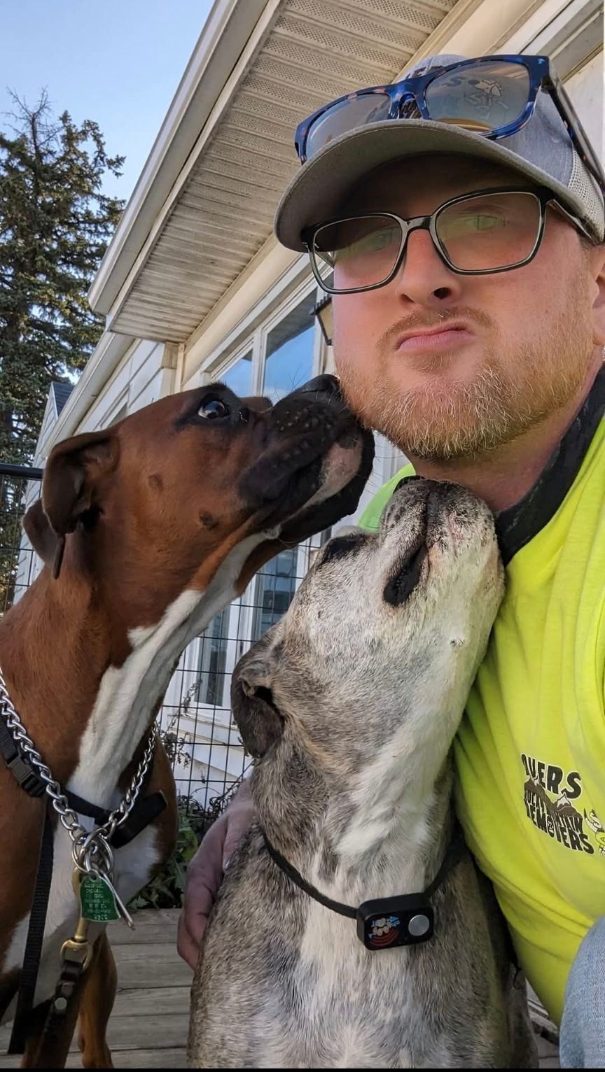 Man with two dogs, one brown and one gray, getting kisses outside. Man wears glasses and a hat.