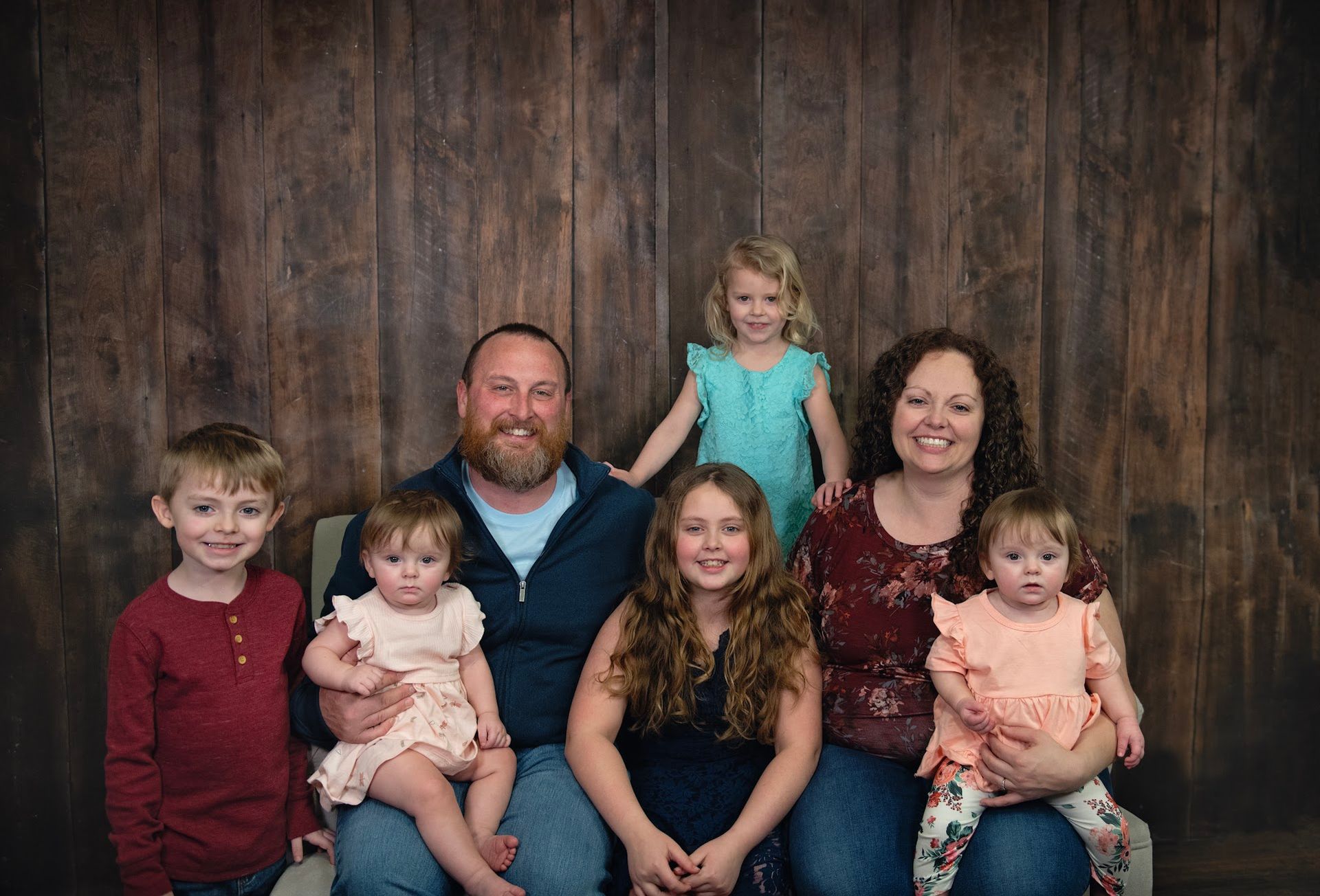 Family portrait in front of a wood backdrop. Six children and two adults smiling.