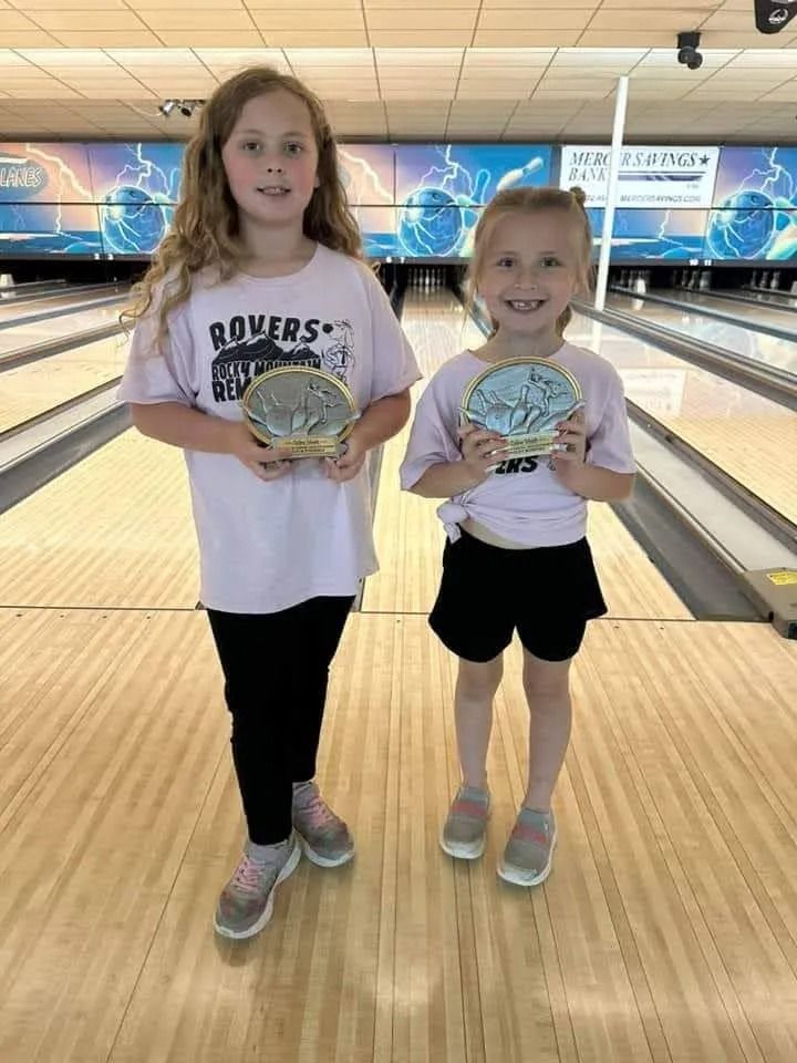 Two young girls at a bowling alley, holding trophies. One wears black pants, the other black shorts. Both smile.