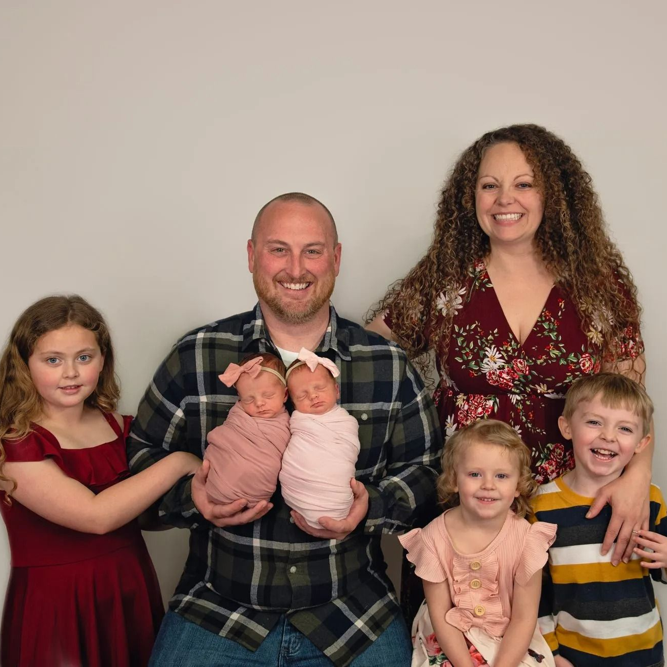 Family of seven poses, holding twin babies. Cheerful expressions. Indoor studio setting.
