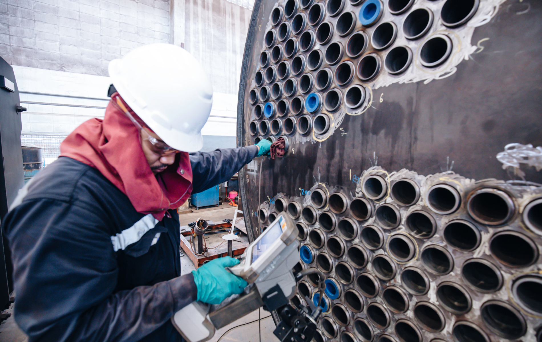 Worker in hard hat inspects metal structure with a device; industrial setting.