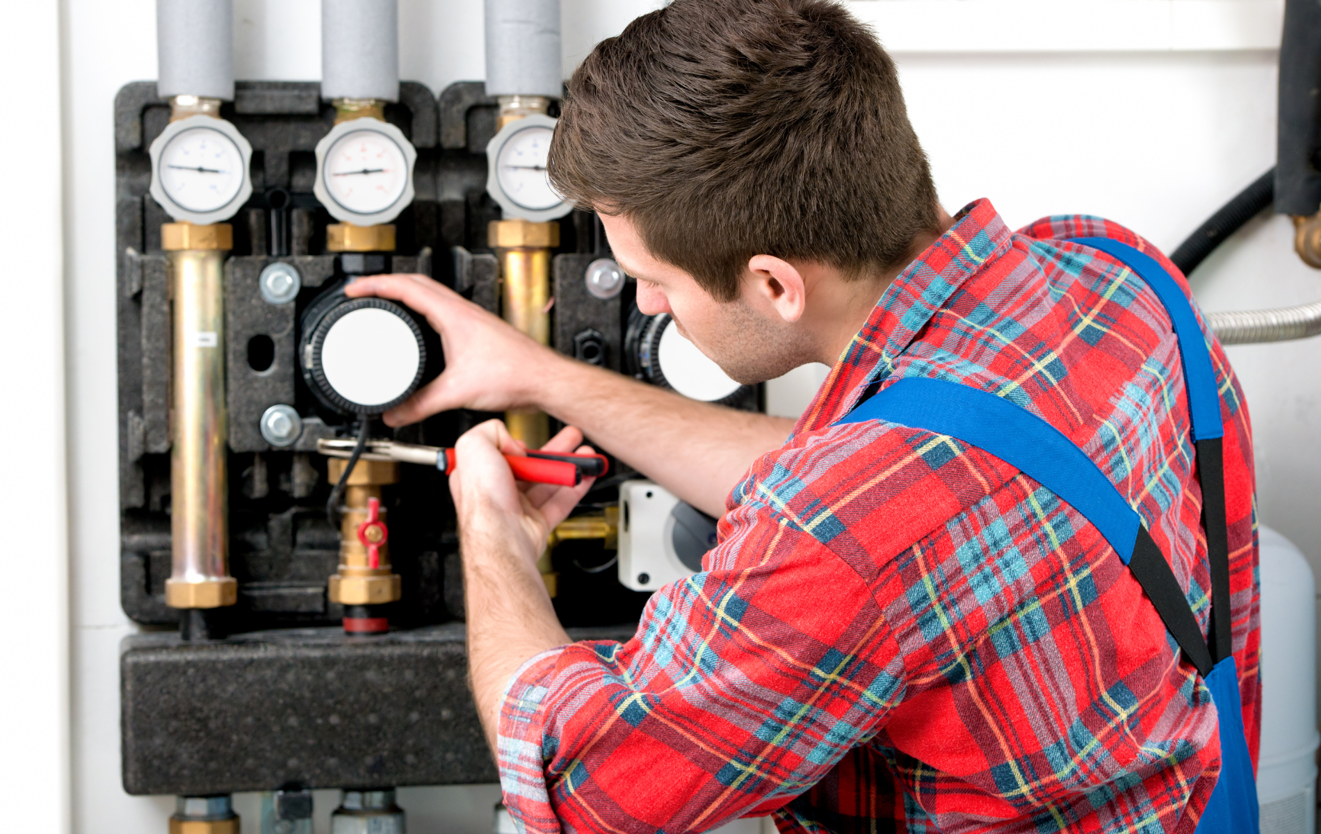 Plumber in red plaid shirt and blue overalls repairs machinery.