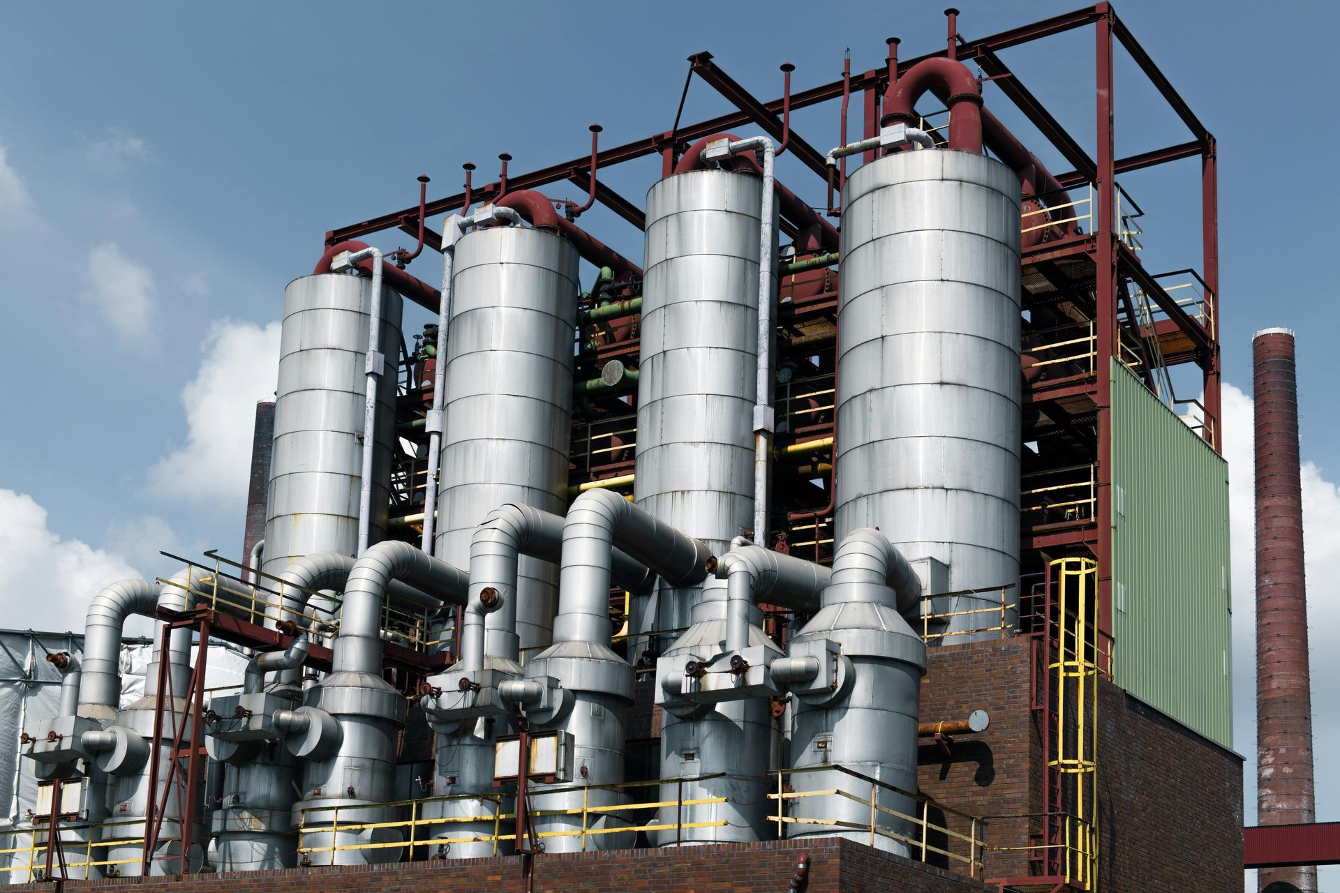 Industrial plant with four large cylindrical tanks, pipes, and a brick smokestack against a blue sky.