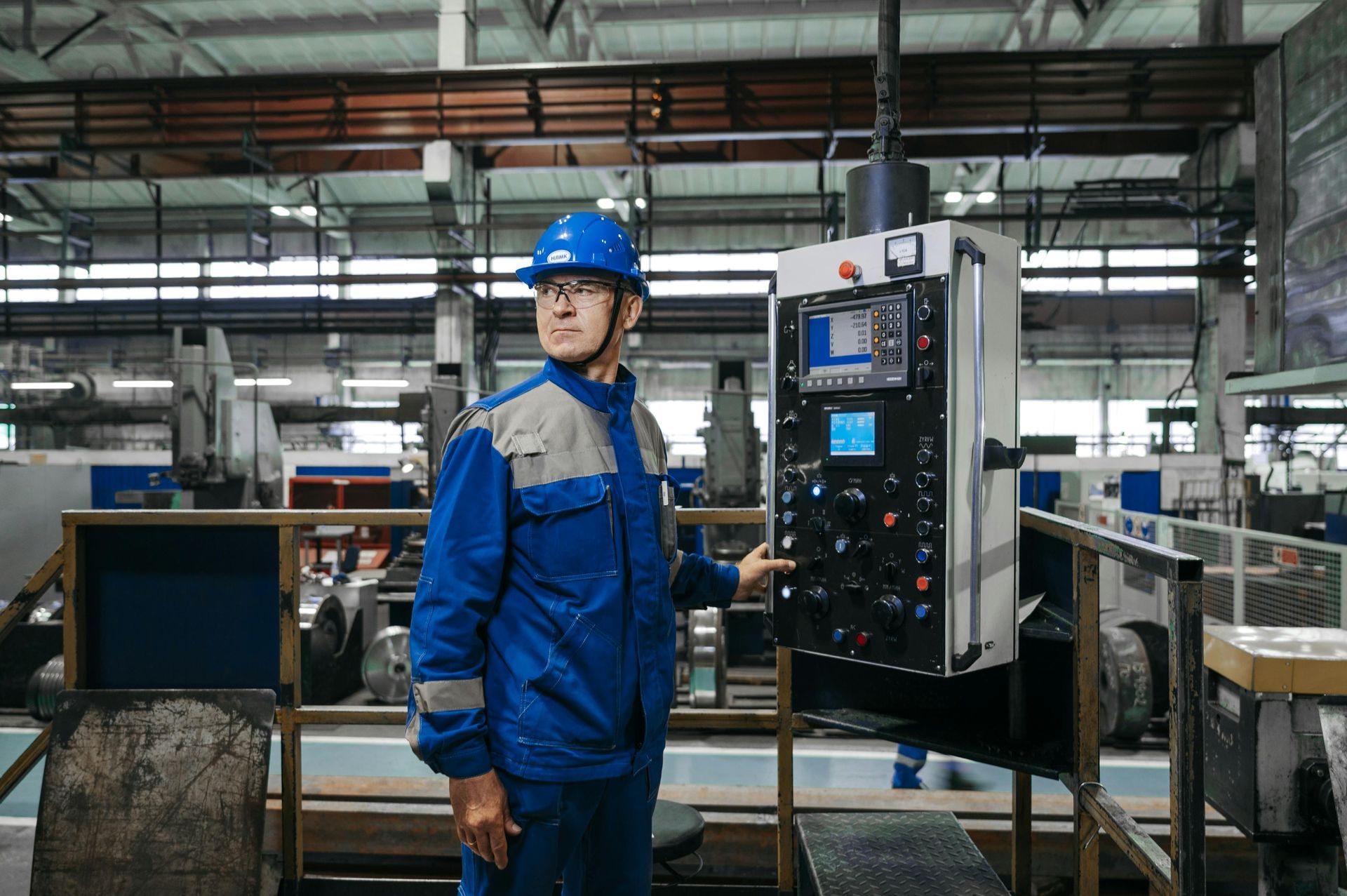 Factory worker in blue uniform and hard hat standing beside a control panel in an industrial workshop
