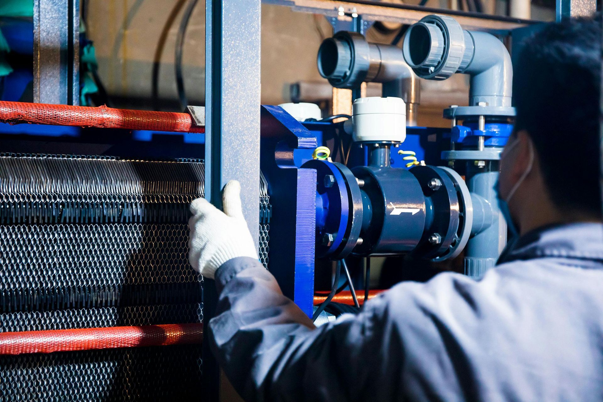 A worker in gloves inspects industrial machinery with pipes and a metal mesh filter in a factory setting.
