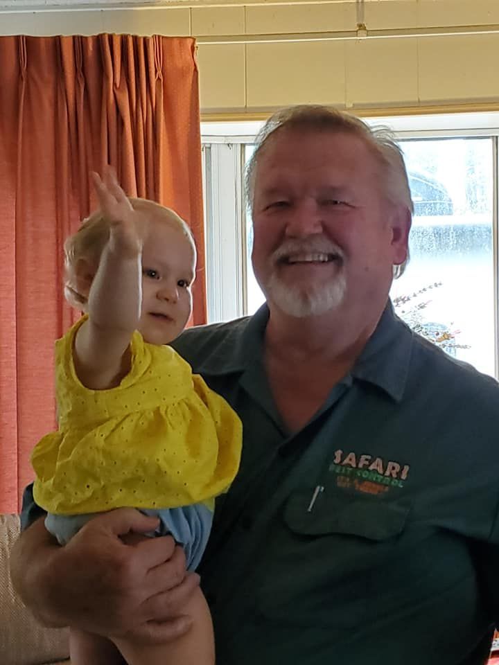 Man in green shirt holds a baby in yellow dress waving a hand. Indoors, smiling.